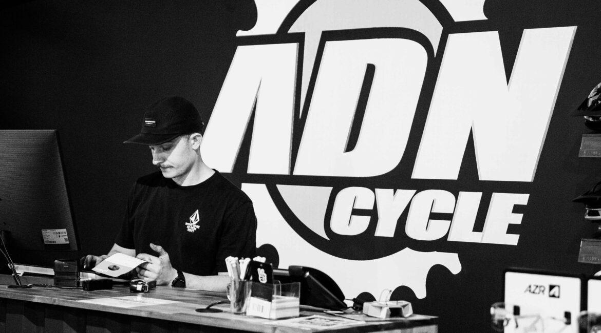 A shop counter with a man in a black cap and shirt handling papers, in front of a large wall logo featuring a gear and bold letters behind him.