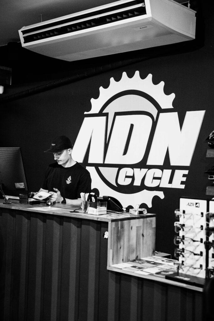 A shop counter with a man in a black cap and shirt handling papers, in front of a large wall logo featuring a gear and bold letters behind him.