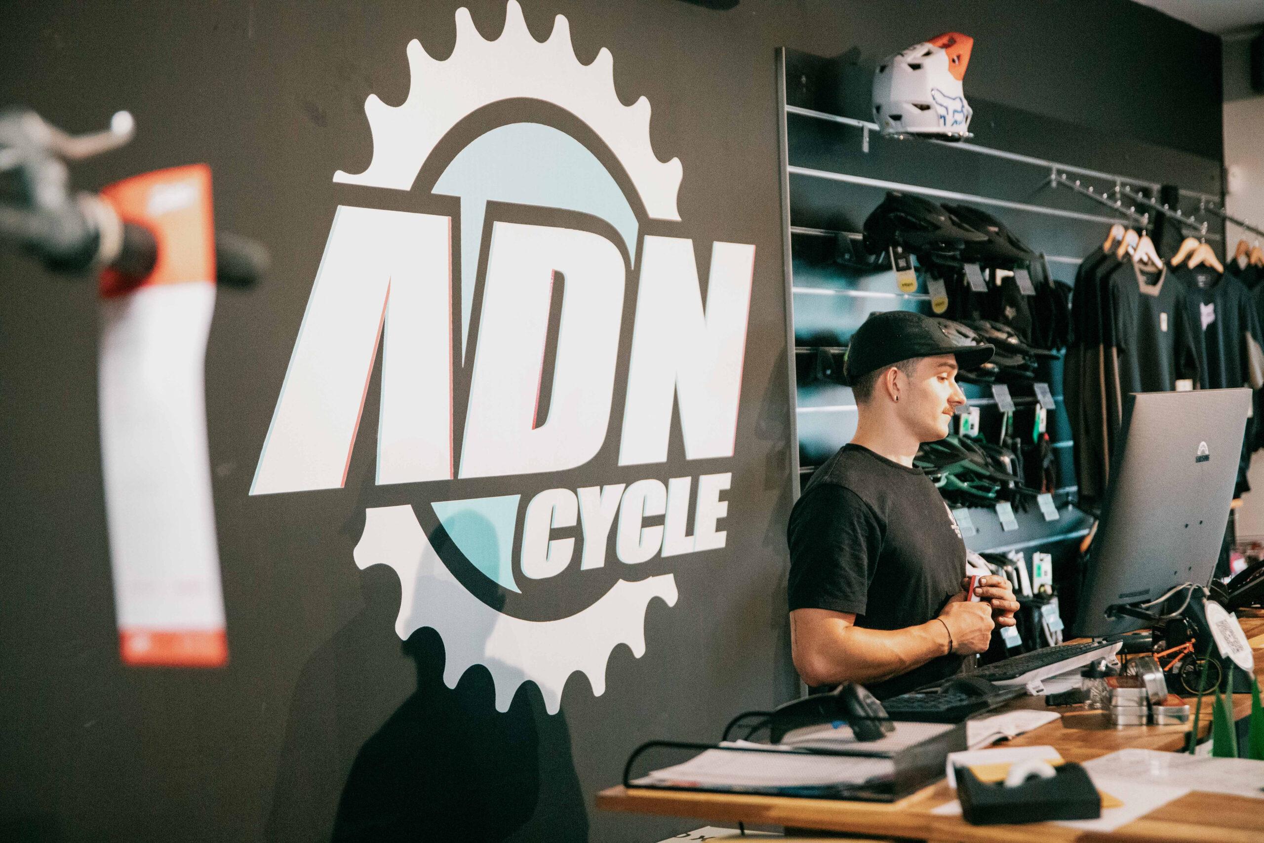 image00057 - ADN Cycle Motorcycle shop counter with staff member in a black cap, wall logo featuring a gear, and helmets on display.