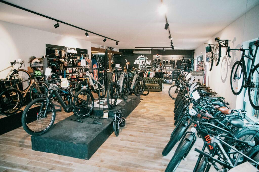 Showroom of a bike shop with numerous bicycles on display, gear on shelves, and customers browsing in a bright space with wooden floors.