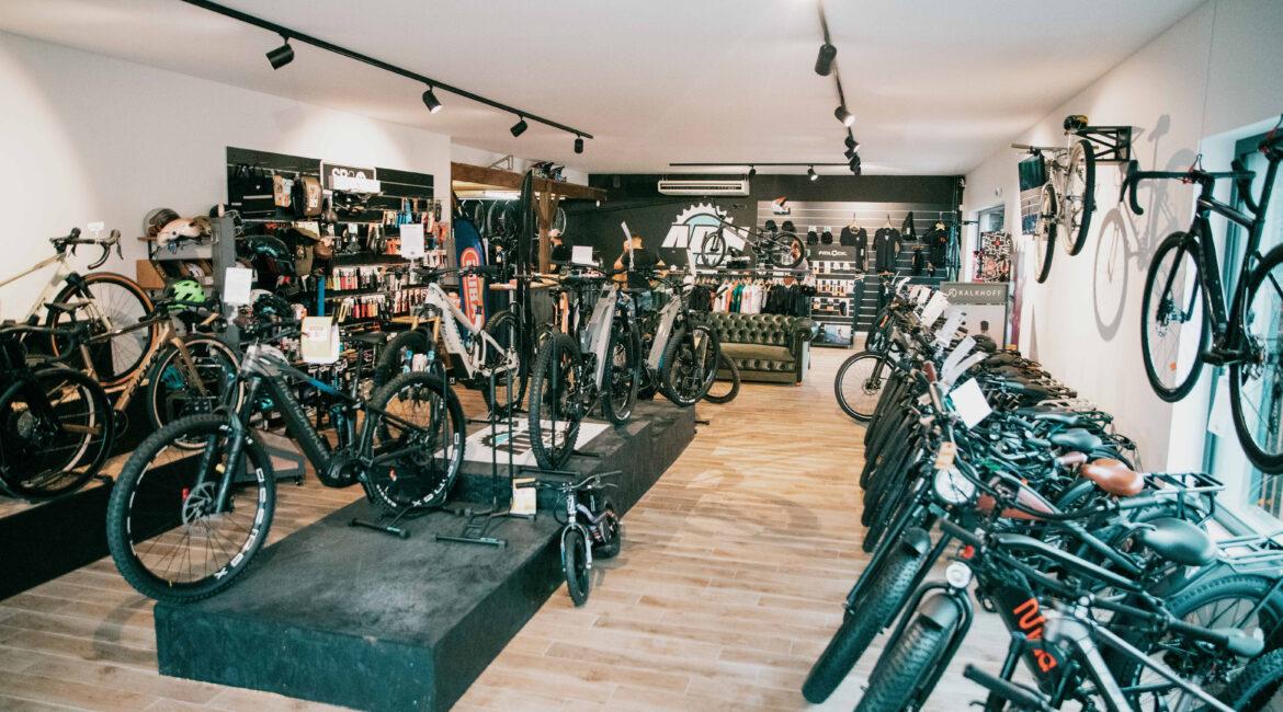 Showroom of a bike shop with numerous bicycles on display, gear on shelves, and customers browsing in a bright space with wooden floors.