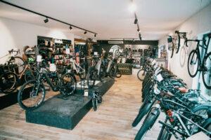 Showroom of a bike shop with numerous bicycles on display, gear on shelves, and customers browsing in a bright space with wooden floors.