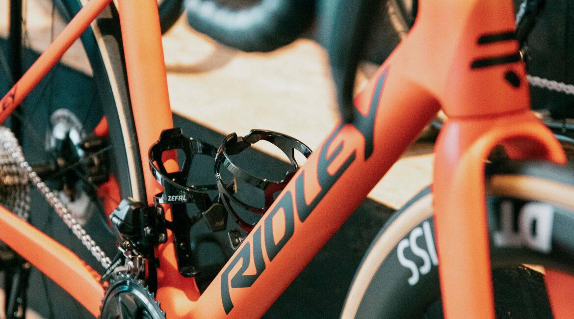 Close-up of an orange bicycle frame with a water bottle cage, showing the chainrings and front fork in view.