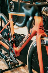 Close-up of an orange bicycle frame with a water bottle cage, showing the chainrings and front fork in view.