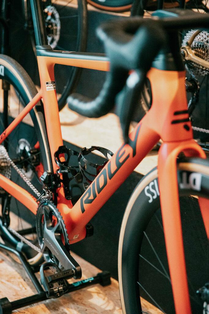 Close-up of an orange bicycle frame with a water bottle cage, showing the chainrings and front fork in view.