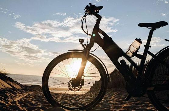 balades-velo-landes-adn - ADN Cycle Electric bike on a sandy beach at sunset, the sun low behind the front wheel creating a silhouette.